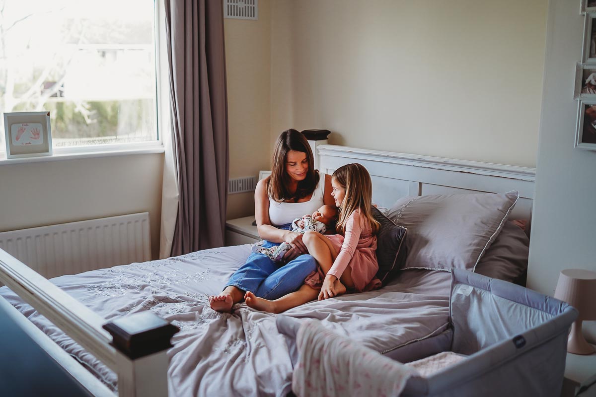 Mum sat on her bed cuddling her two daughters during her newborn photoshoot in Newbury