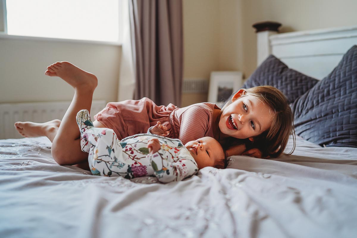 Two Newbury sisters smiling whilst led on the bed during a newborn lifestyle photoshoot