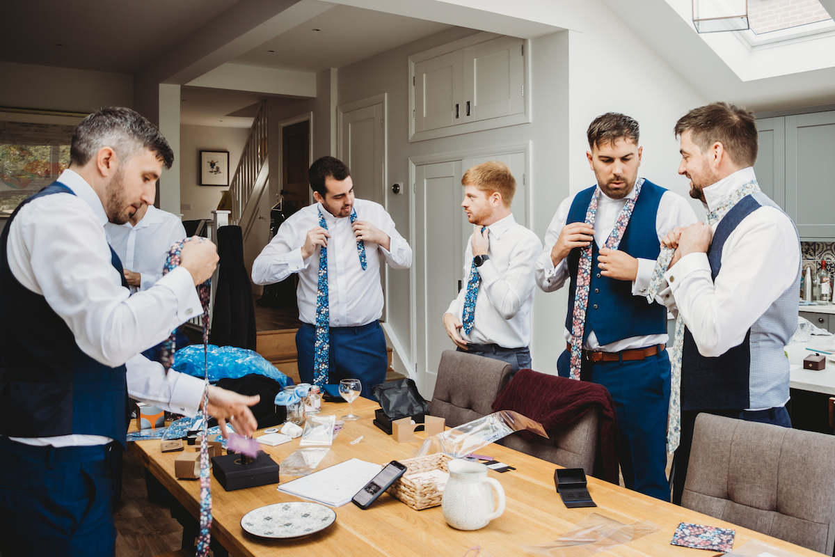 grooms men all helping each other with their ties as they get ready for a wedding