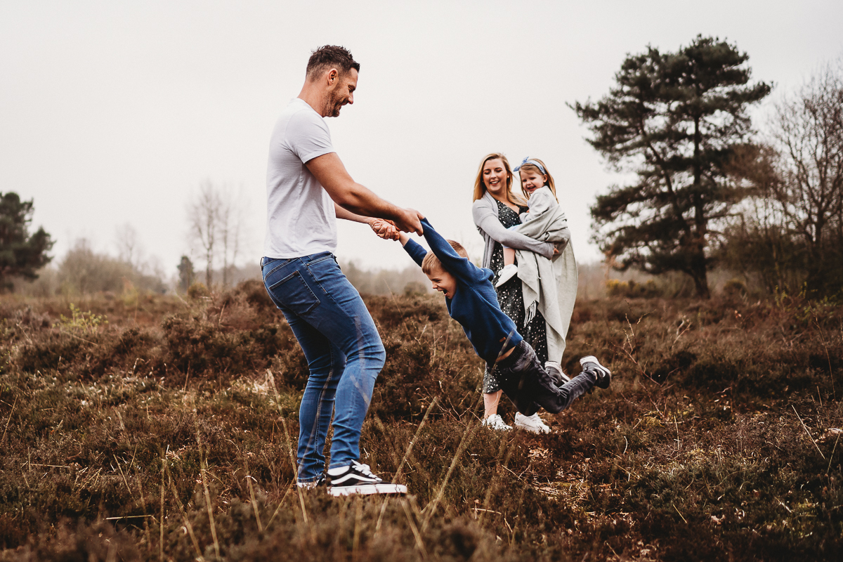 Mum and daughter looking on as dad swings his boy around for a natural family photoshoot