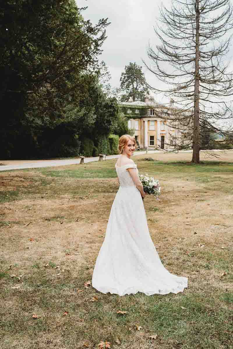 a bride looking over her shoulder with woolley park estate in the background before her woodland ceremony