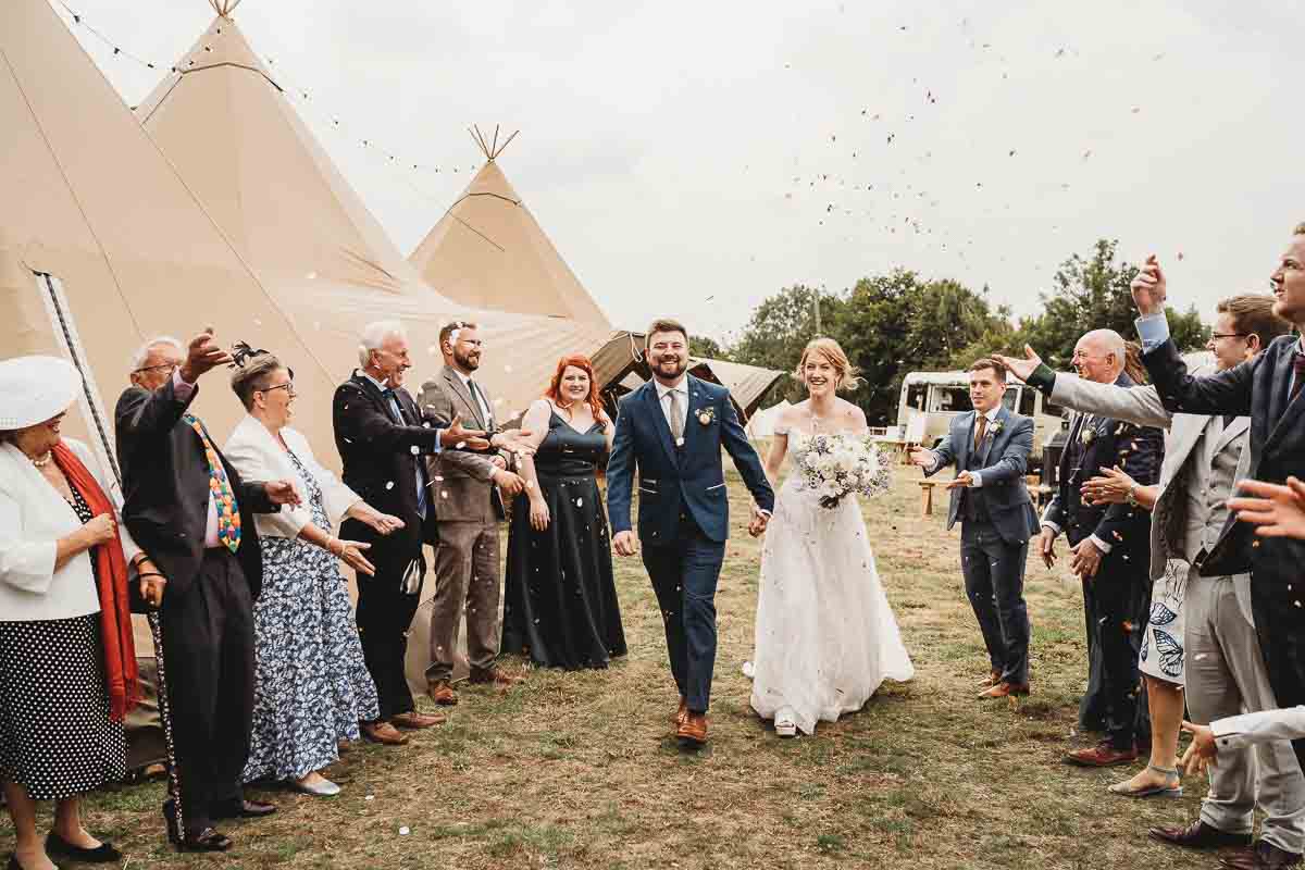 a bride and groom walking through a confetti line up in front of the tipis for a tipi wedding