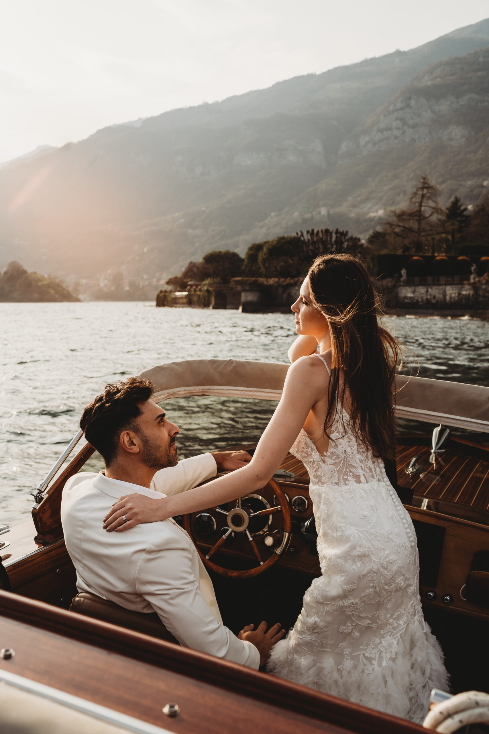 a bride leaning over to chat to her husband as he steers the boat across lake como