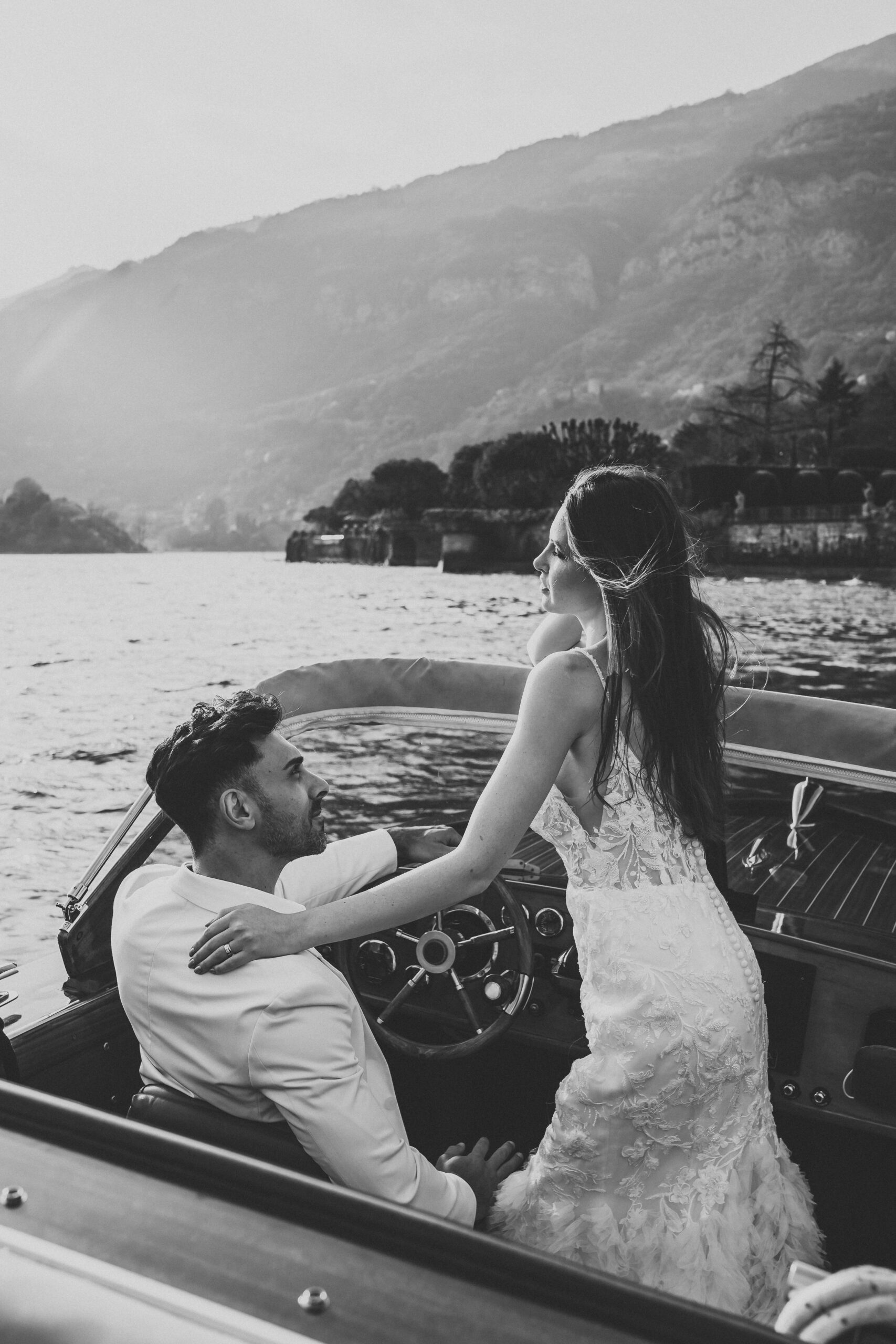 a bride leaning towards her husband as he steers their boat across lake como