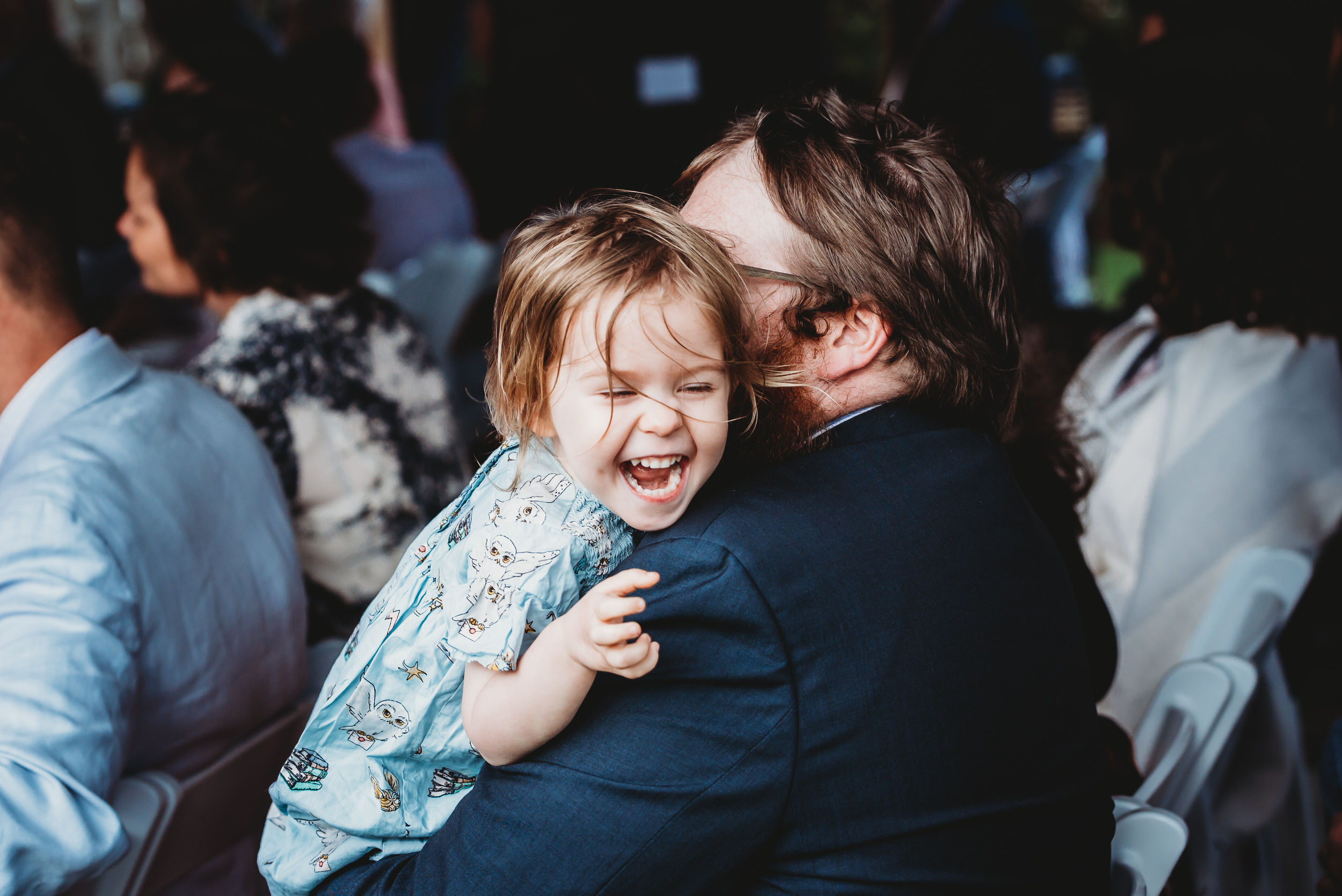 a little girl laughing whilst looking over her fathers sholder during a wedding