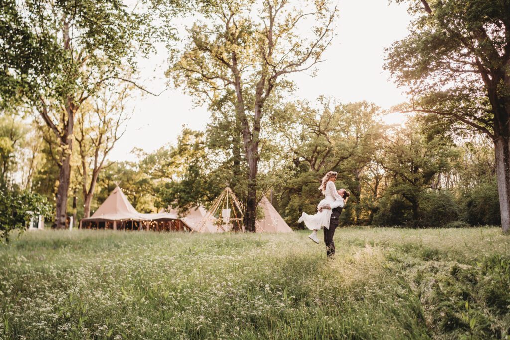 a groom lifts his wife up for a kiss in the long grasses just after their festival wedding 