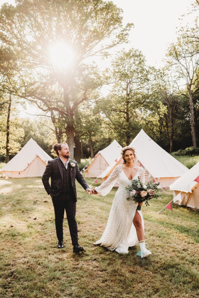 a bride and groom walking hand in hand through bell tents in a festival field 