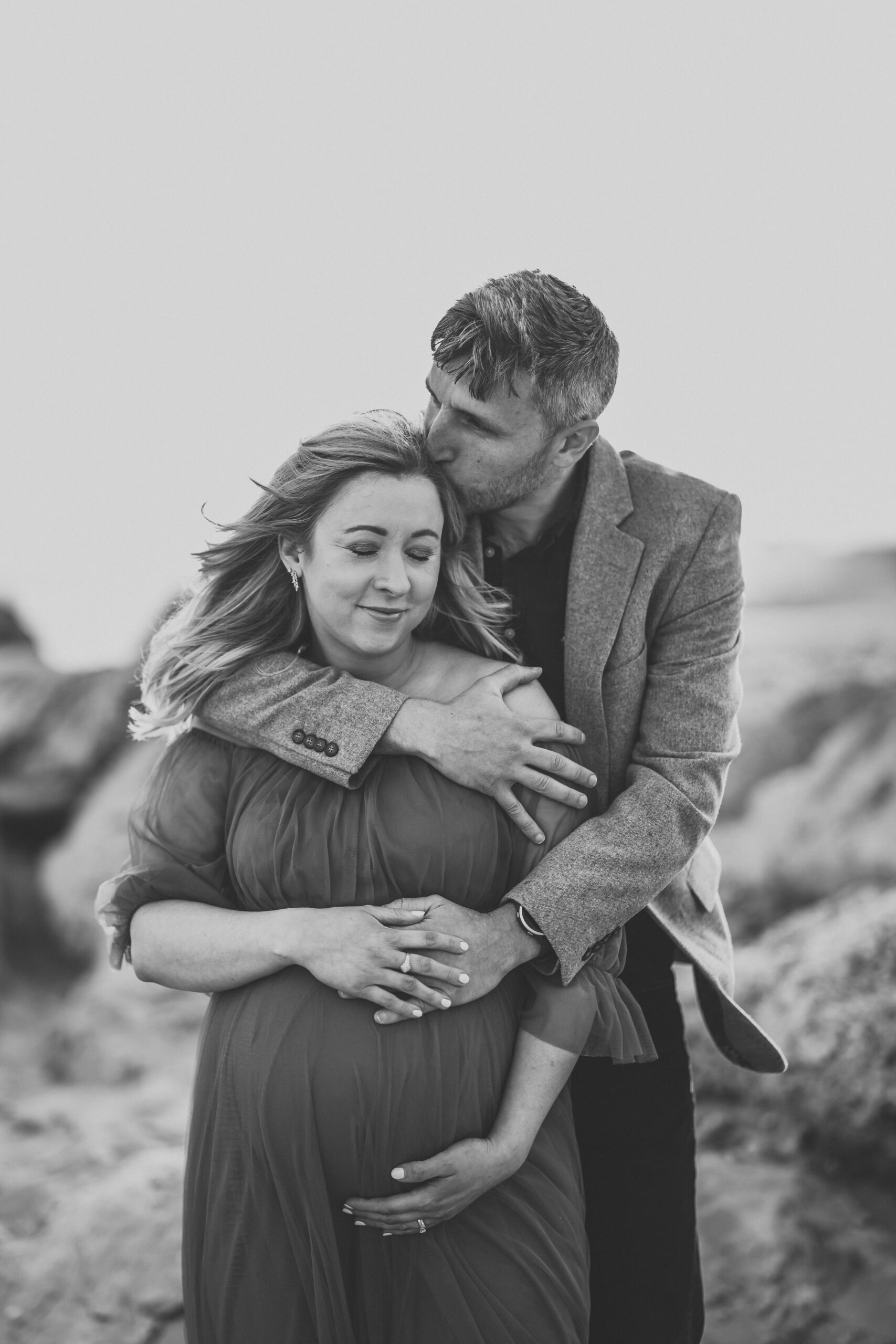 a husband cuddling into his pregnant wife from behind during a beach maternity session