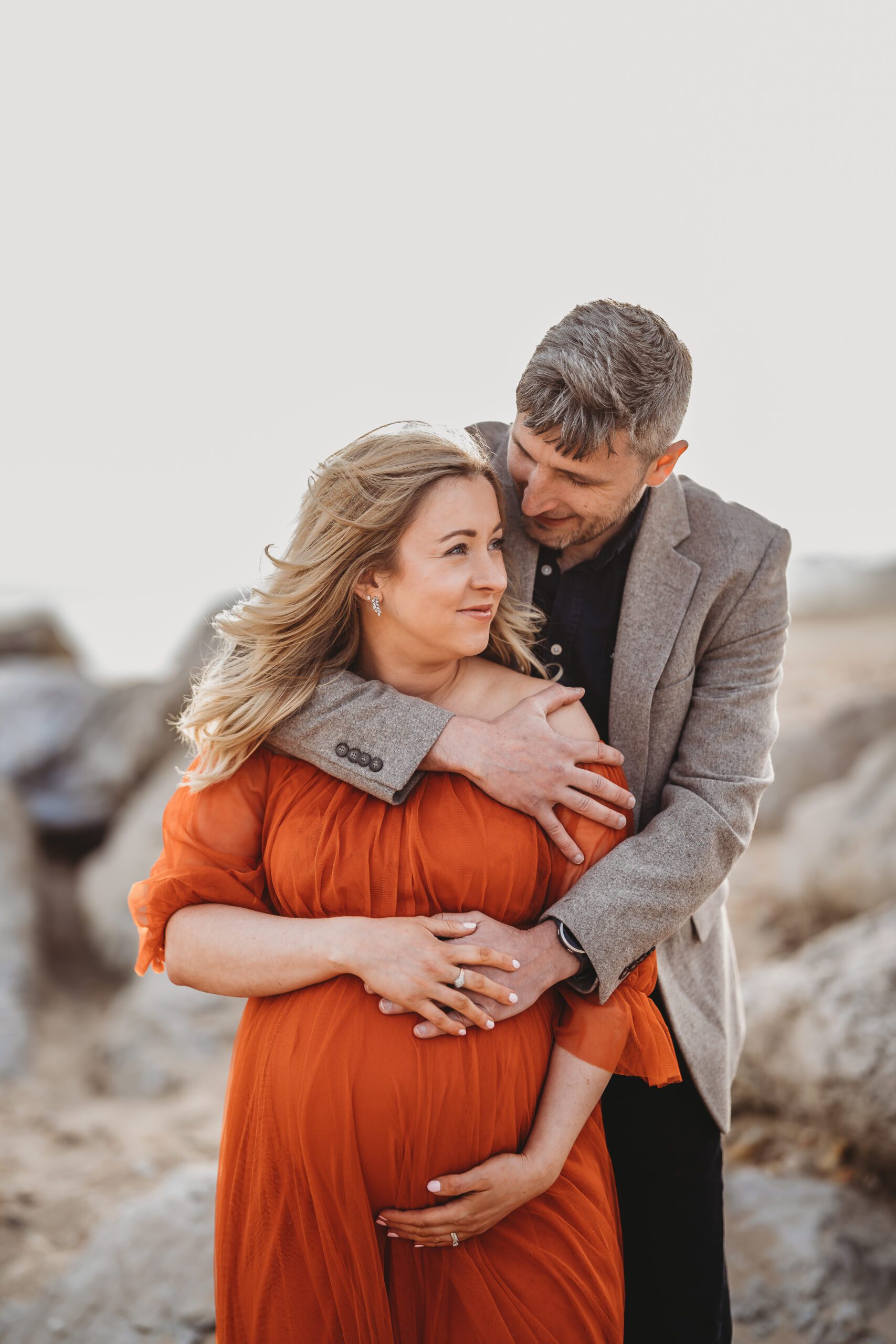 a husband cuddling into his pregnant wife from behind during a beach maternity session