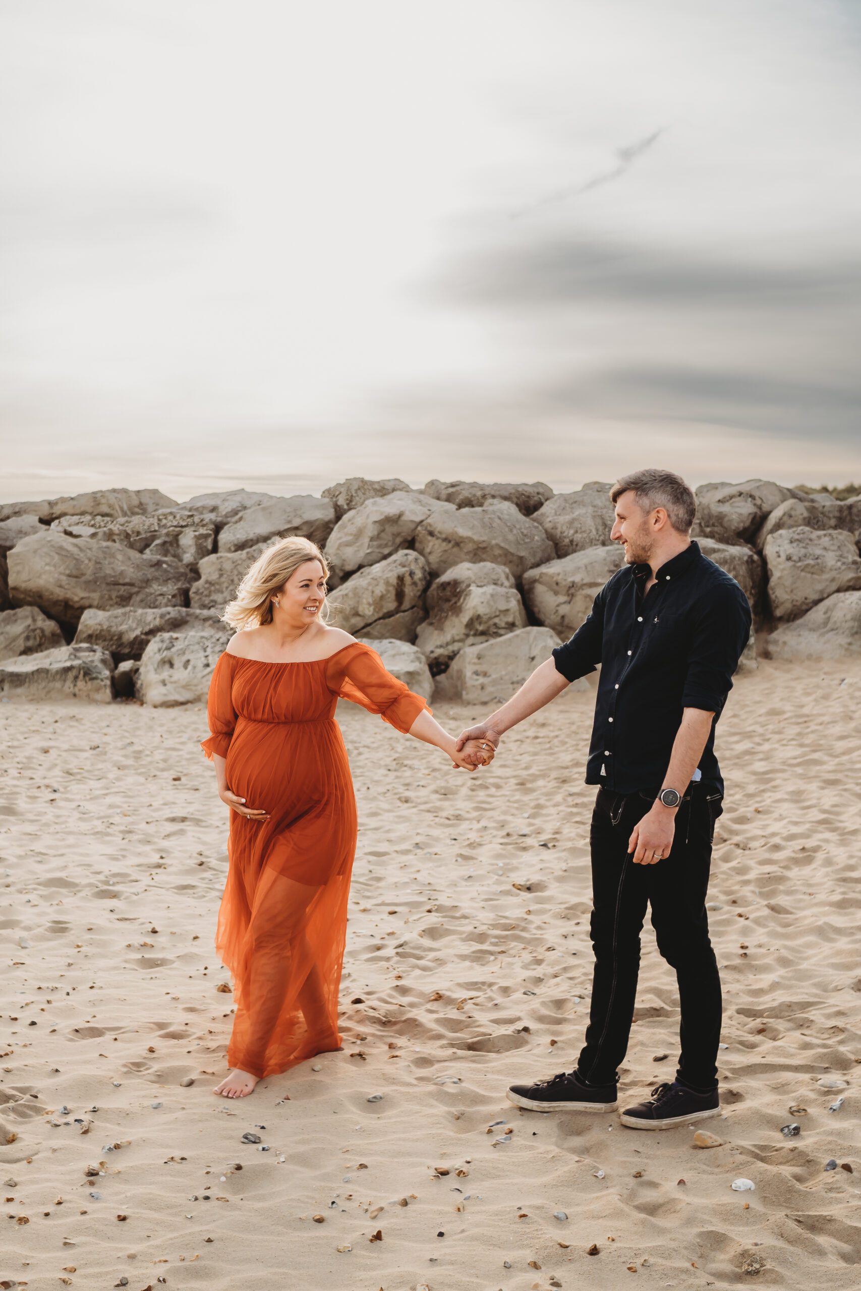 a husband and pregnant wife walking along a beach holding hands 