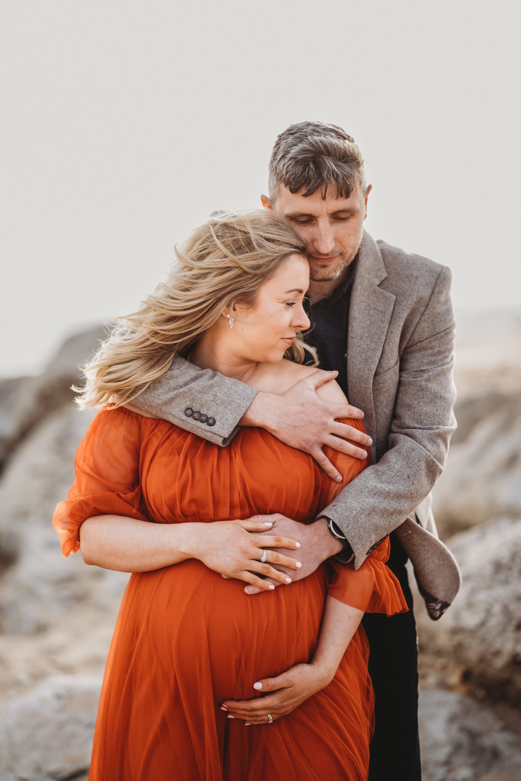 a husband cuddling into his pregnant wife from behind during a beach maternity session