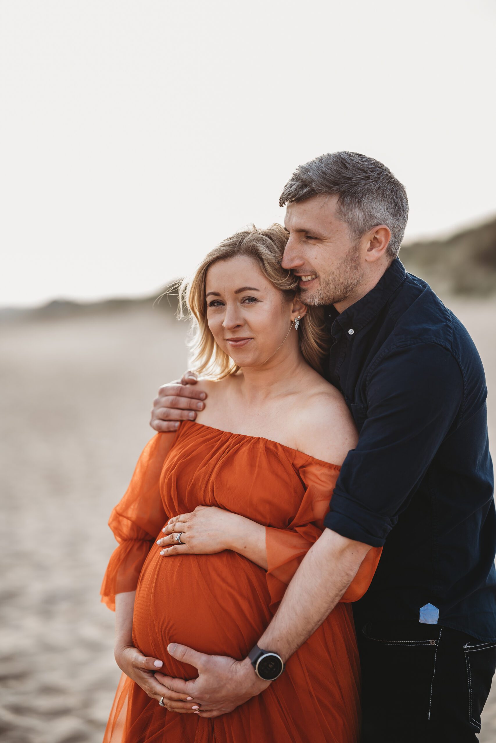 a husband cuddling his pregnant wife from behind during a beach maternity session taken by a newbury maternity photographer