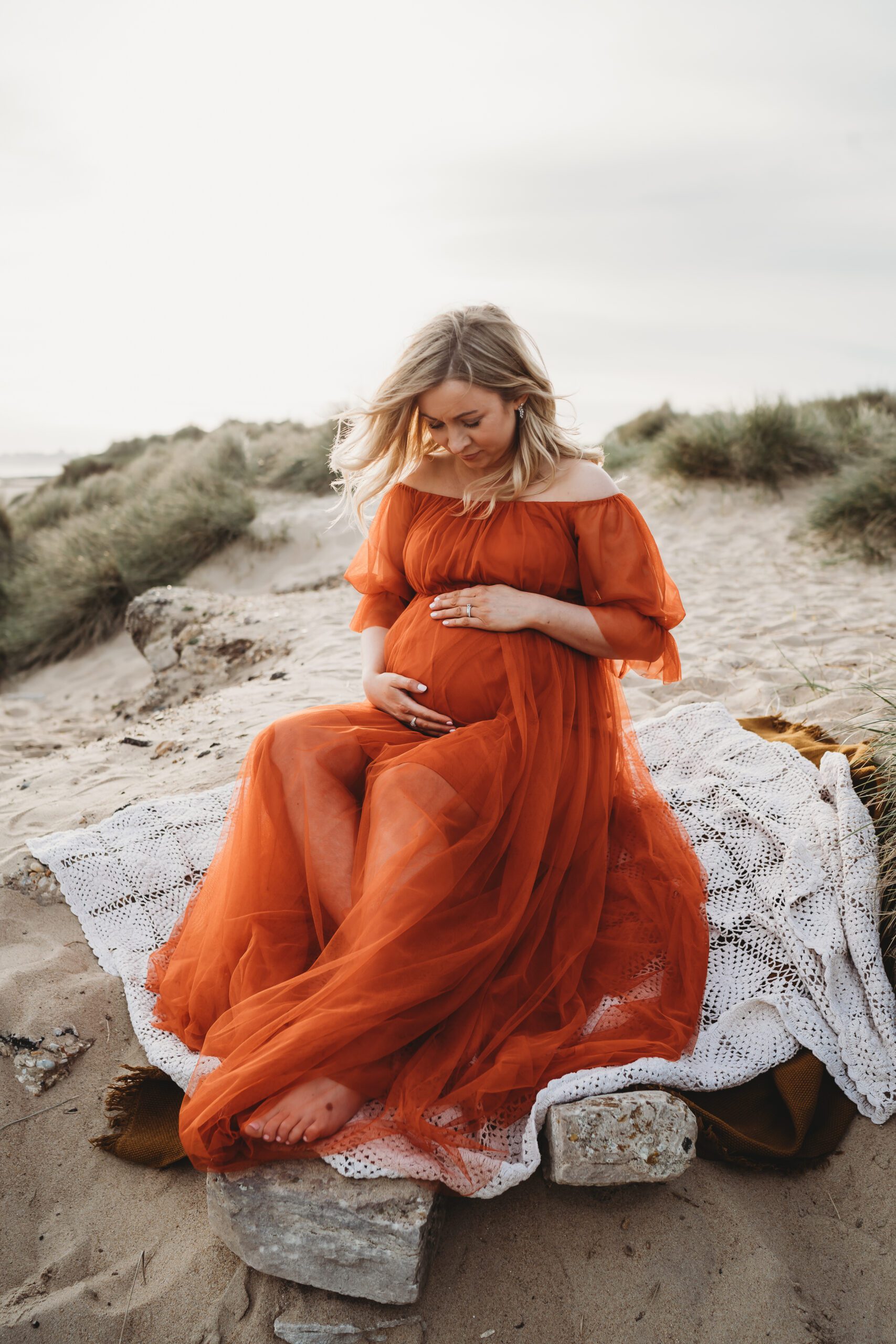 a pregnant lady sitting on a rock holding on to her bump in a bright orange dress taken by a newbury maternity photographer