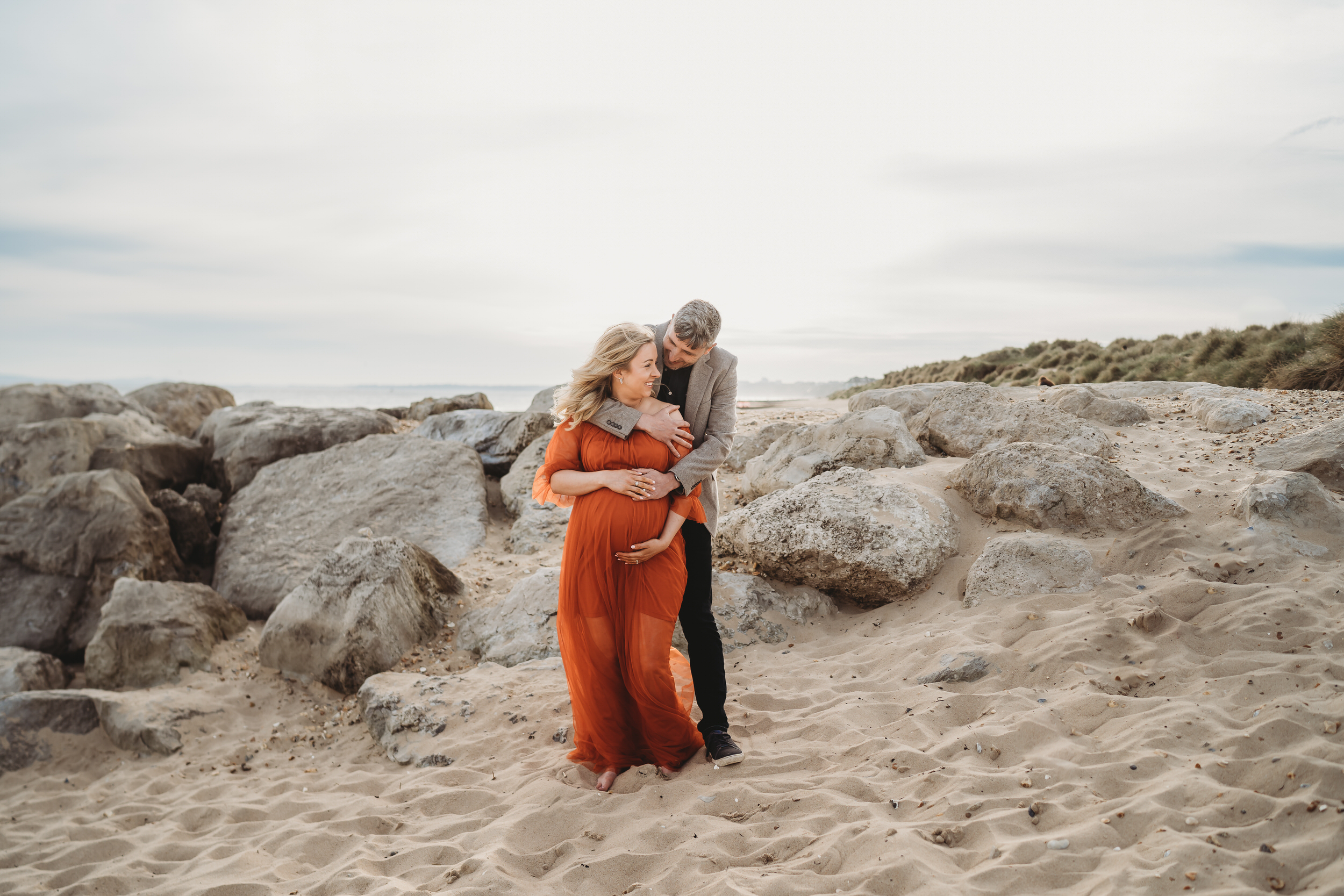 a husband cuddling into his pregnant wife from behind during a beach maternity session