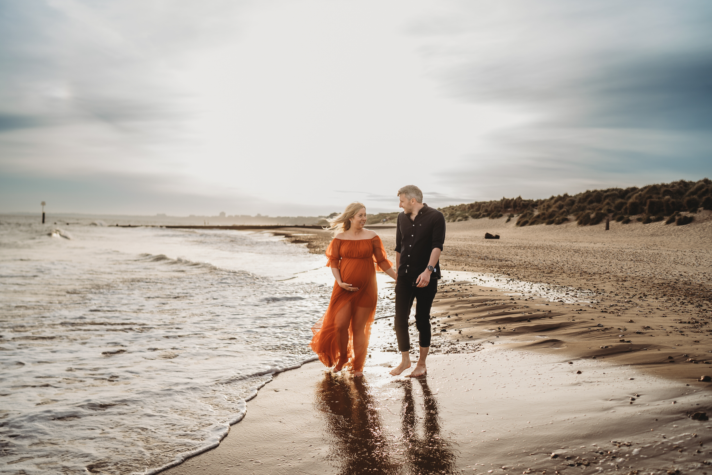 a husband and pregnant wife hold hands as they walk through the waves at Hengistbury Head Beach for a newbury wedding photographer