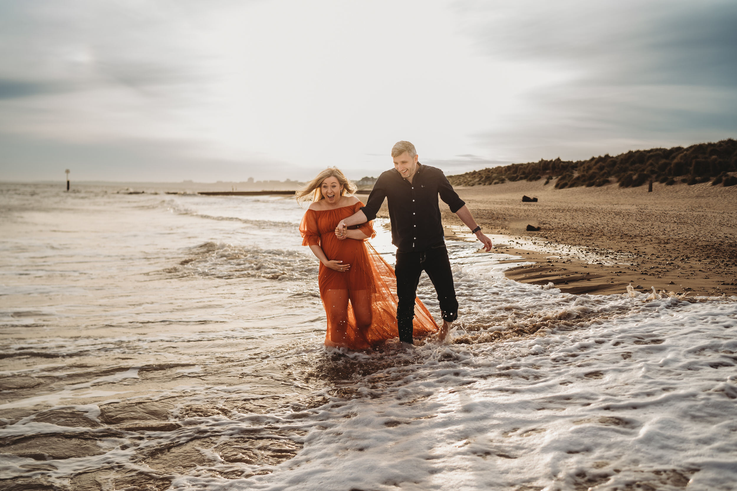 a husband and pregnant wife hold hands and laugh as they walk through the waves at Hengistbury Head Beach for a newbury wedding photographer