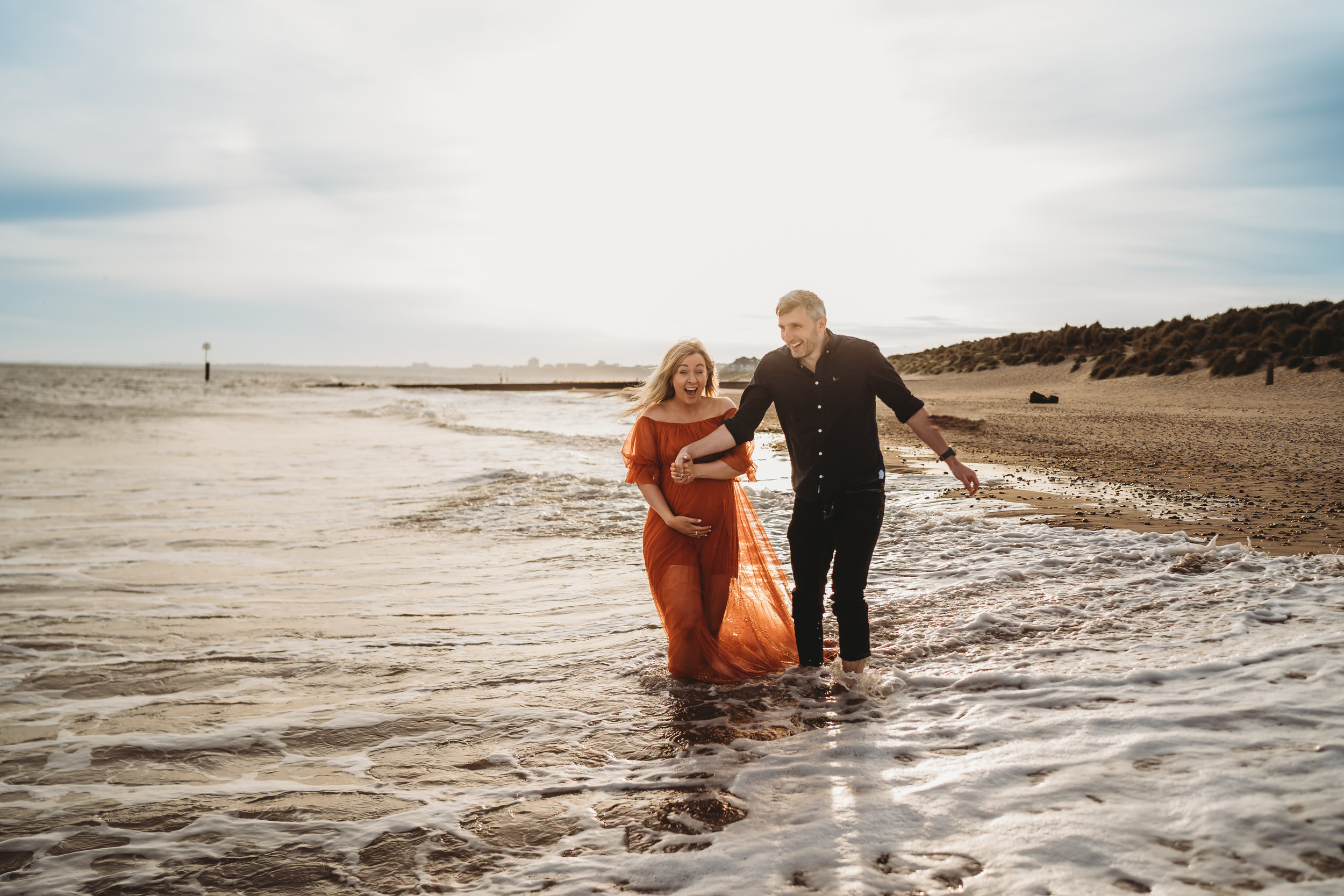 a husband and pregnant wife laugh and hold hands as they walk through the waves at Hengistbury Head Beach for a newbury wedding photographer