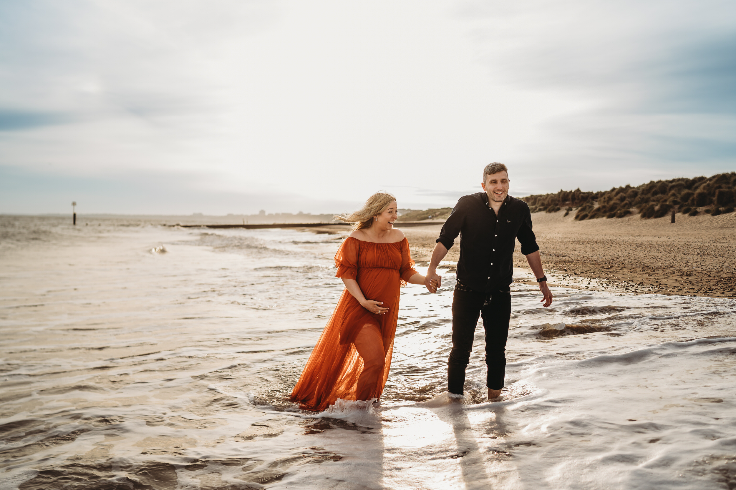 a husband and pregnant wife hold hands as they walk through the waves at Hengistbury Head Beach for a newbury wedding photographer