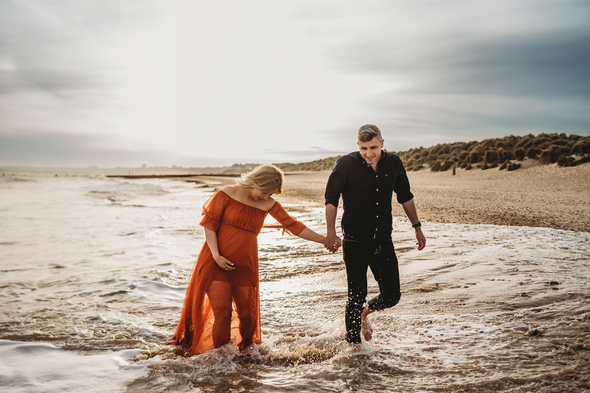 a husband and pregnant wife hold hands as they walk through the waves at Hengistbury Head Beach for a newbury wedding photographer