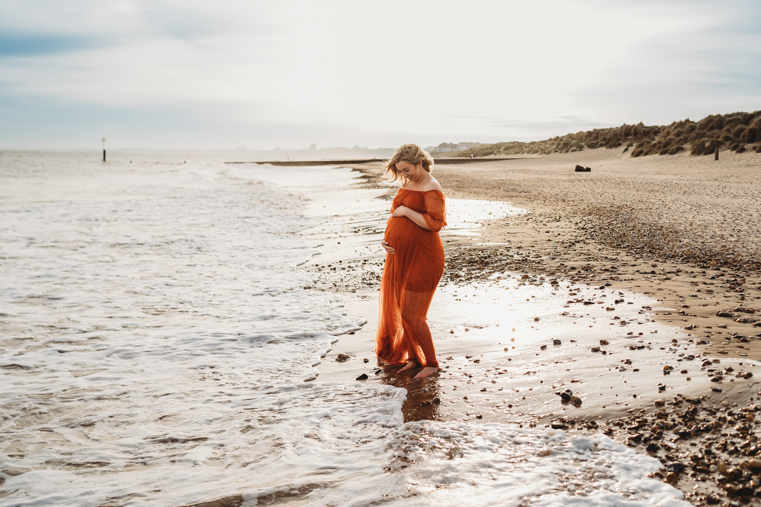 a pregnant lady in a bright orange dress hugging her belly for a maternity beach session for a newbury photographer