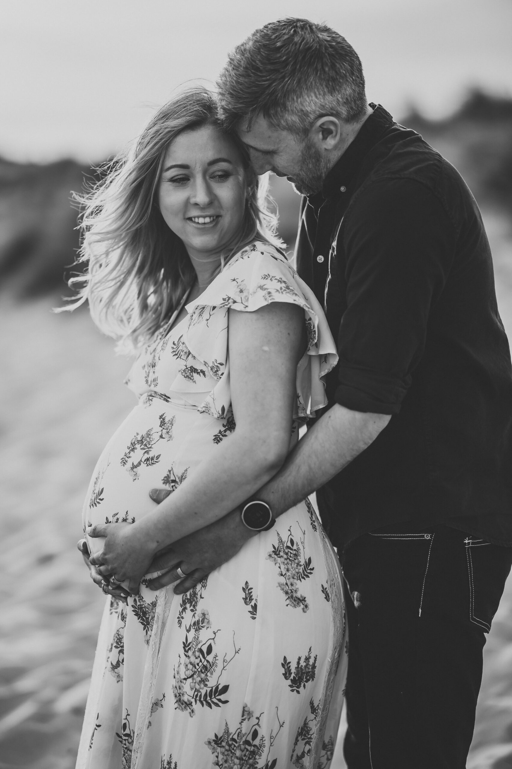 a husband cuddles his wife from behind and her pregnant stomach at hengistbury head beach for a newbury materniry photographer