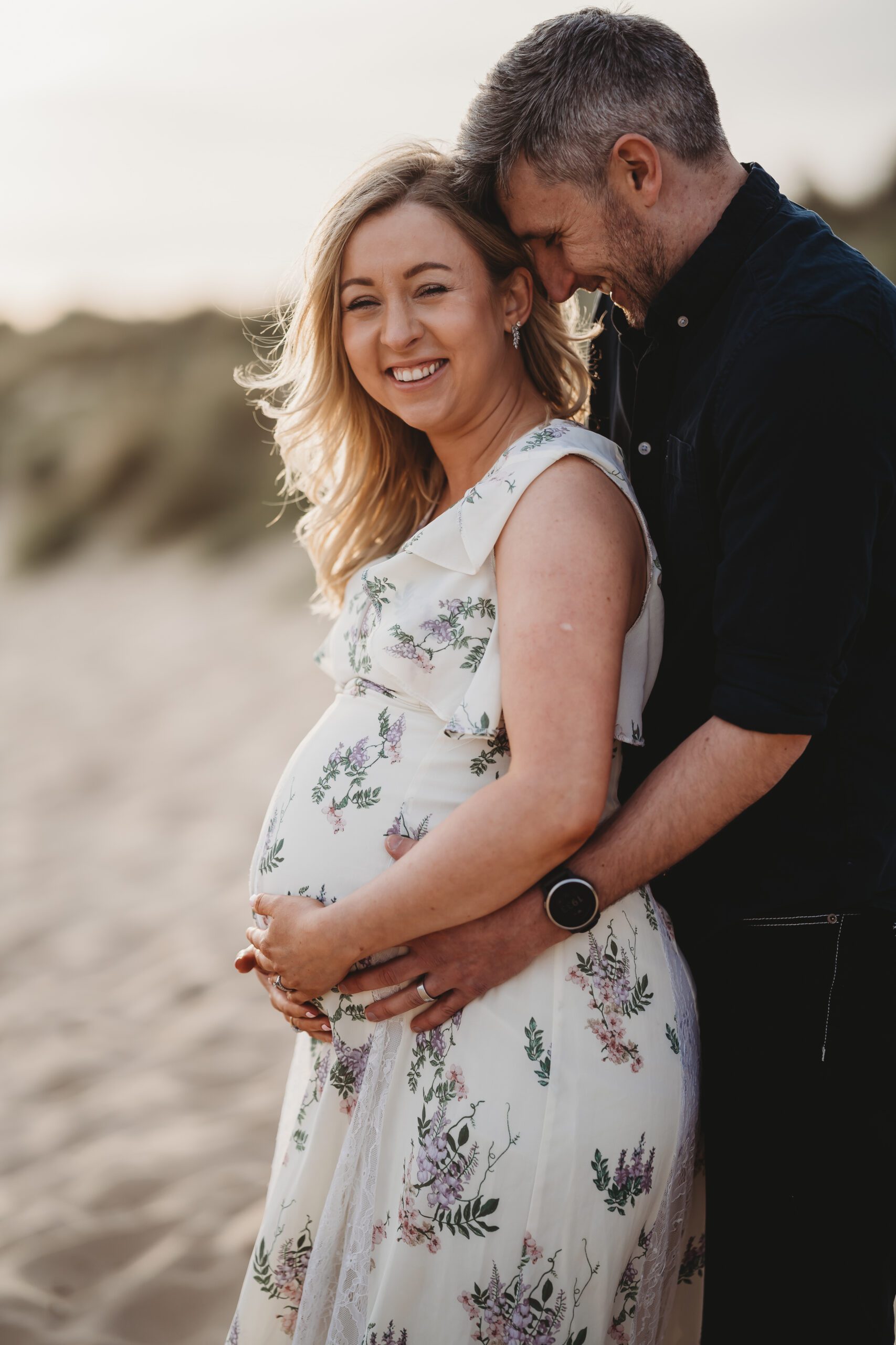 a husband cuddles his wife from behind and her pregnant stomach at hengistbury head beach