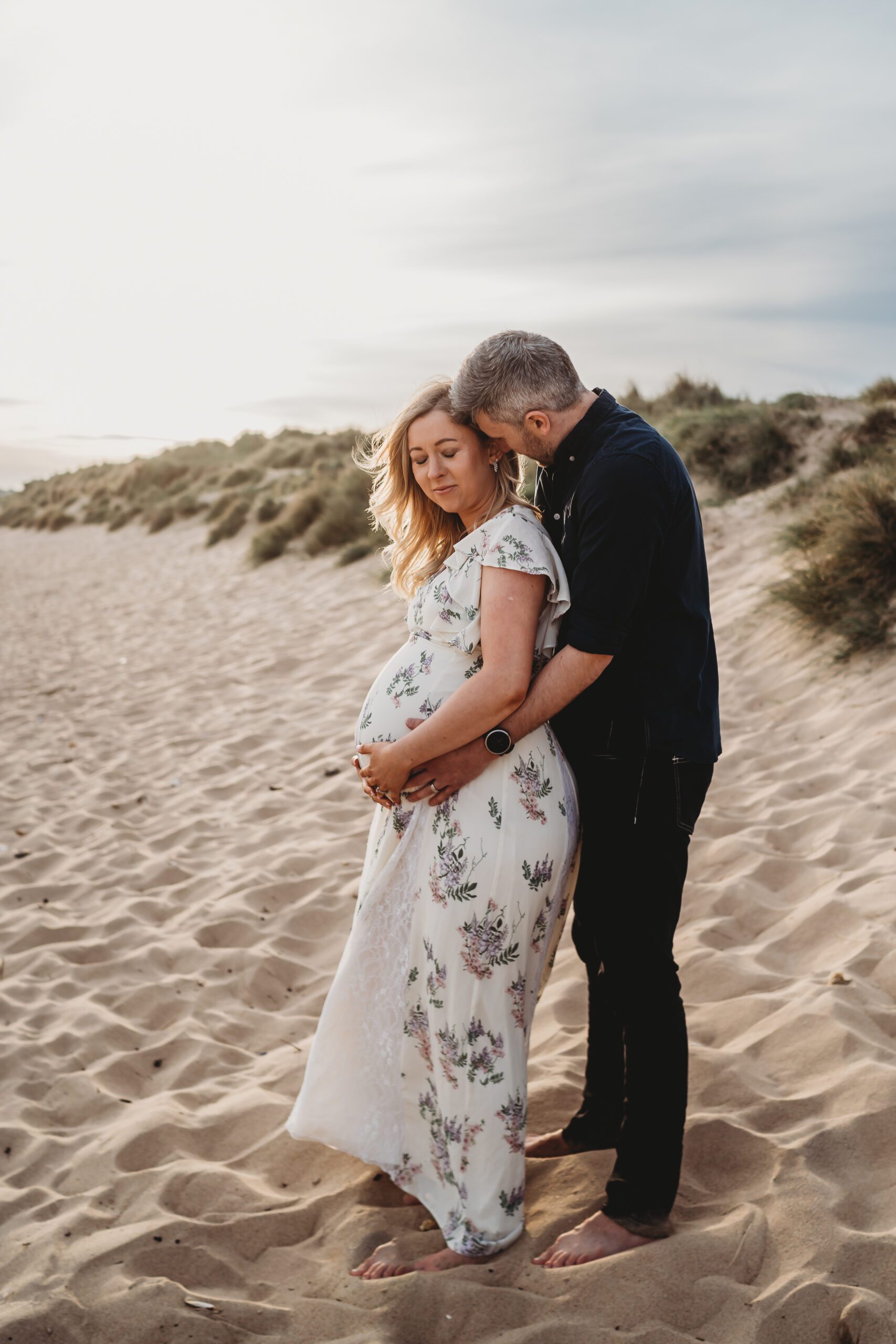 a husband cuddles his wife from behind and her pregnant stomach at hengistbury head beach