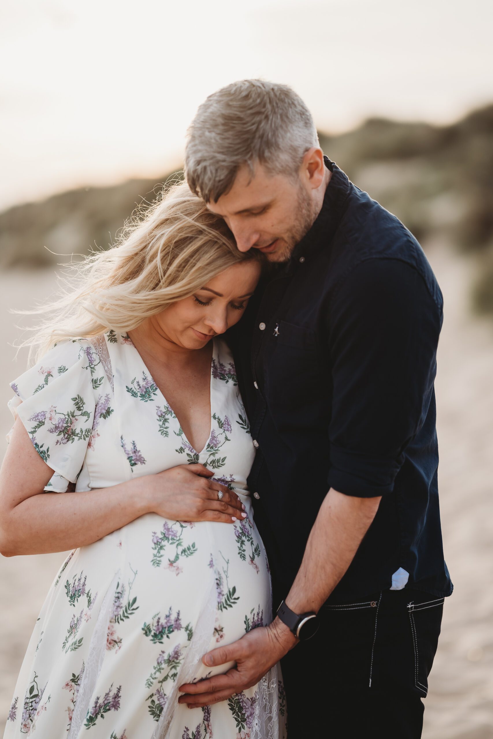 a husband and pregnant wife looking down at her bump as they cuddle on Hengistbury Head Beach