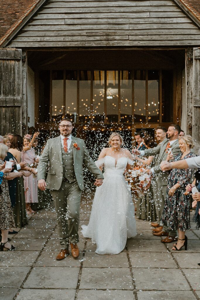 a bride and groom laughing as they walk down a confetti line up outside the barn at Ufton Court in Newbury