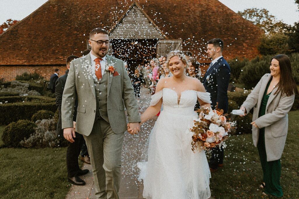 a bride and groom laughing as they walk down a confetti line up outside the barn at Ufton Court in Newbury