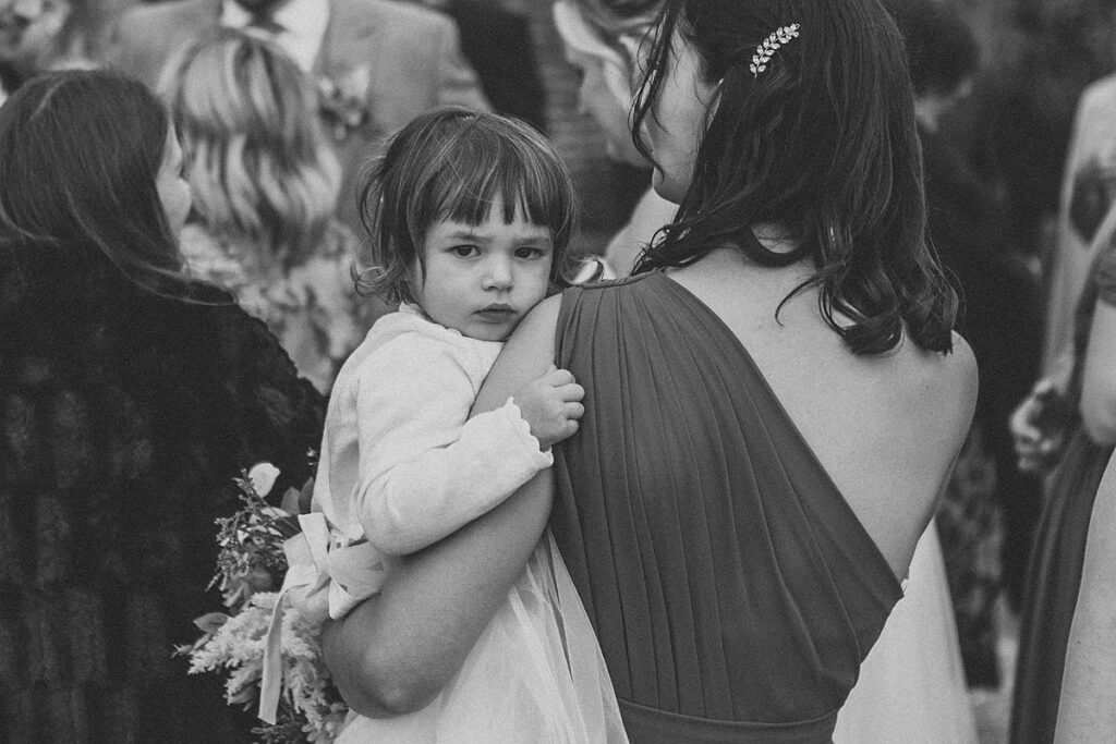 a black and white photo of a flower girl looking over her mums shoulder