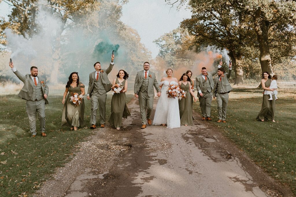 a wedding party walking down a driveway with white, orange and green smoke grenades