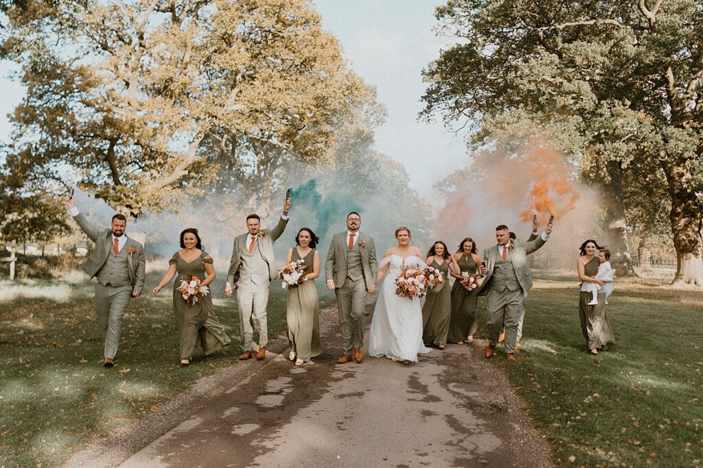 a wedding party walking down a driveway with white, orange and green smoke grenades at Ufton Court