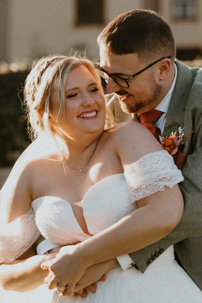 a groom hugging on to his new wife from behind as he nozzles in and laughs