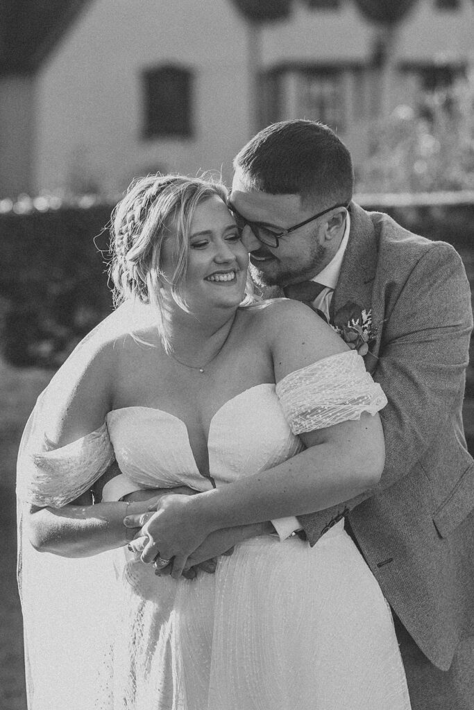 a black and white of a groom hugging on to his new wife from behind as he nozzles in and laughs