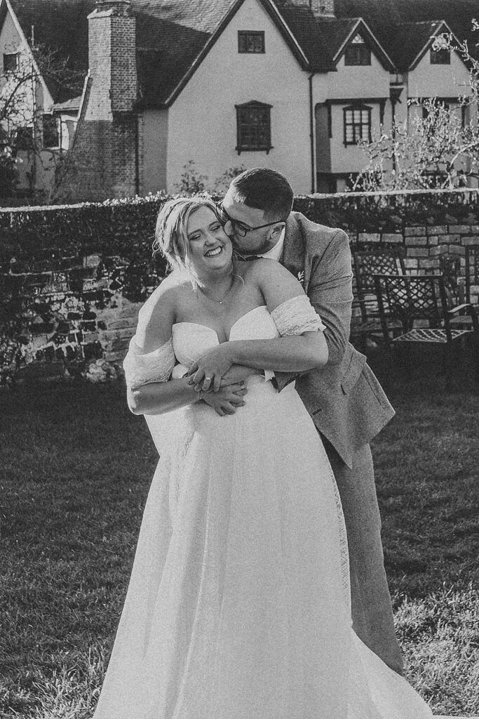 a black and white of a groom hugging on to his new wife from behind as he nozzles in and laughs