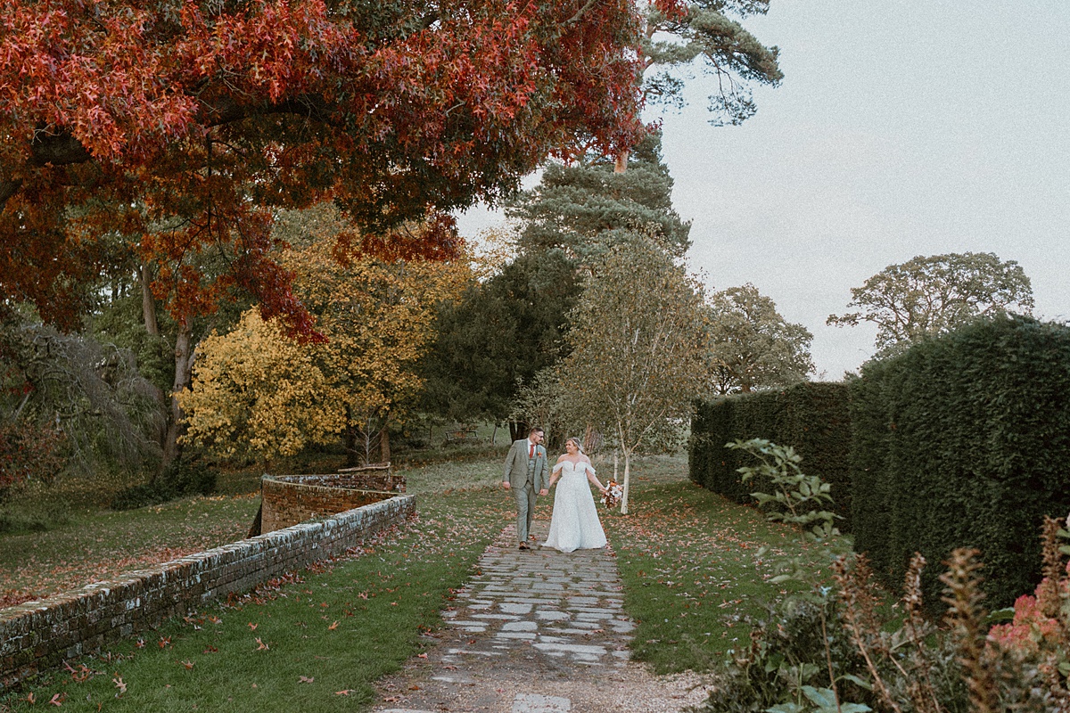 a bride and groom walking along a pathway at Ufton Court surrounded by colourful autumnal trees