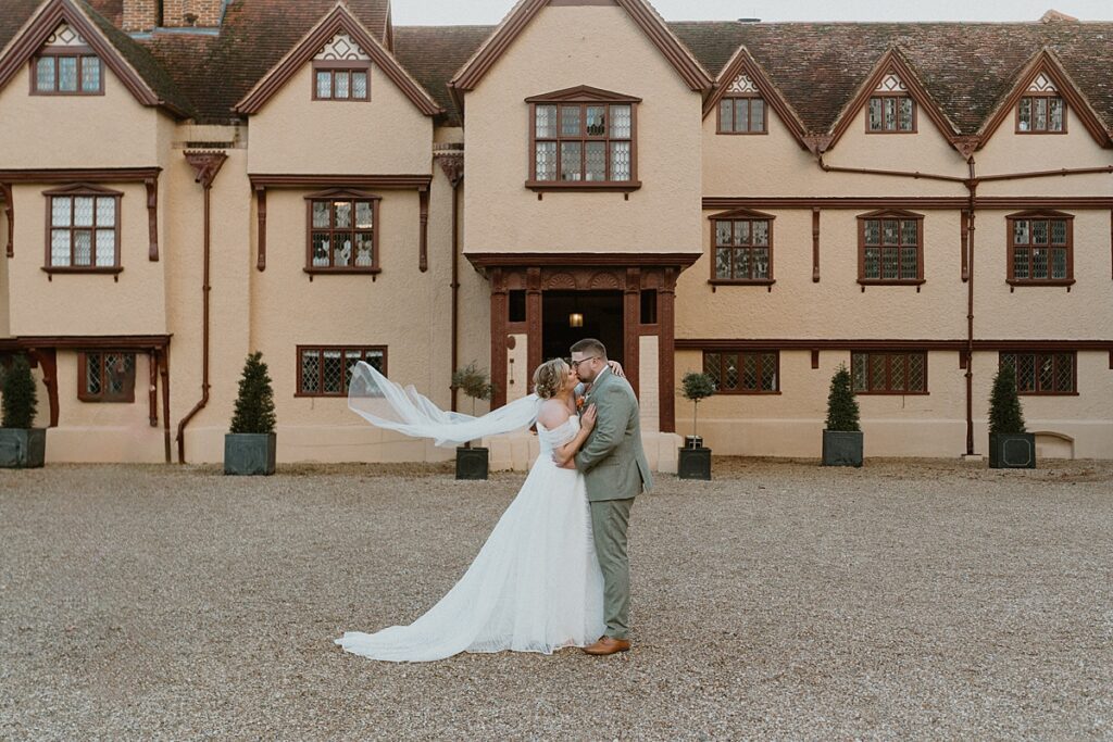 a bride and groom cuddling as her veil lifts with the wind in front of Upton Courts manor house