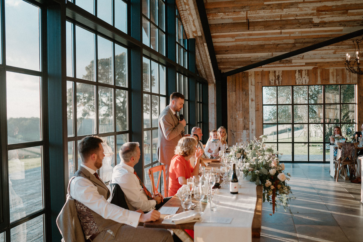 a groom stands as he gives his wedding speech in front of huge barn windows