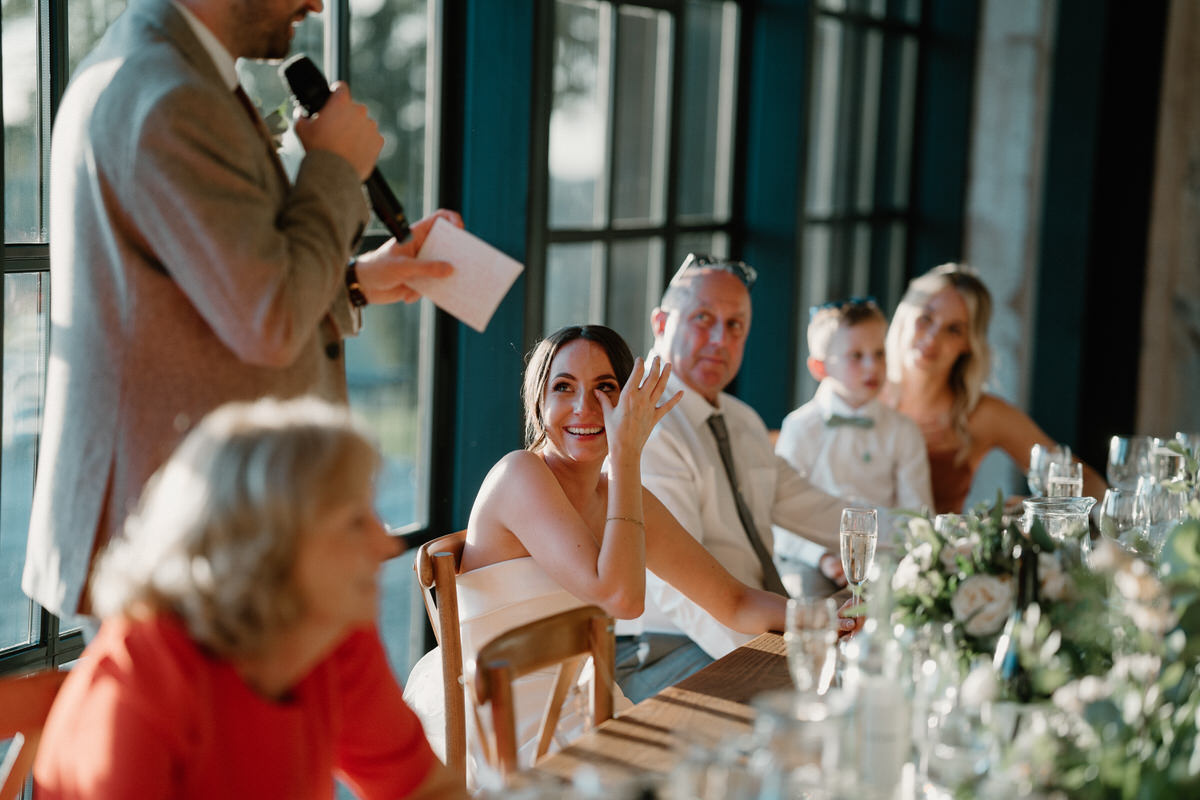 a bride dabs away a tear as she smiles and watches her new husband give his speech