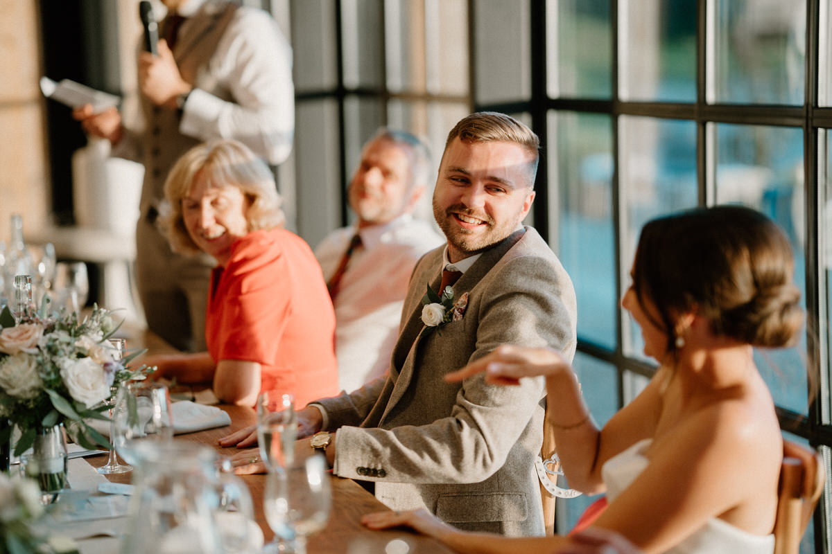 a bride and groom smile and look at each other whilst sat at the top table