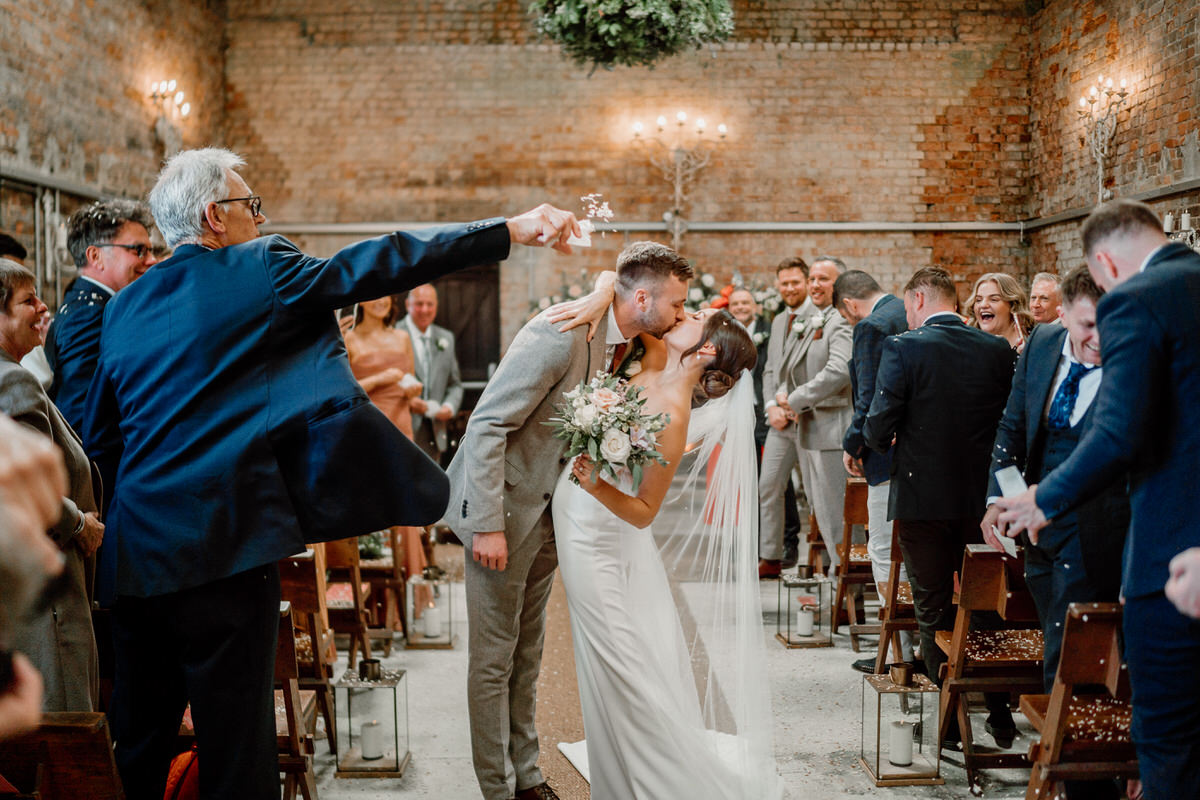 a bride and groom stop for a kiss half way back down the aisle after getting married in a luxury barn in surrey