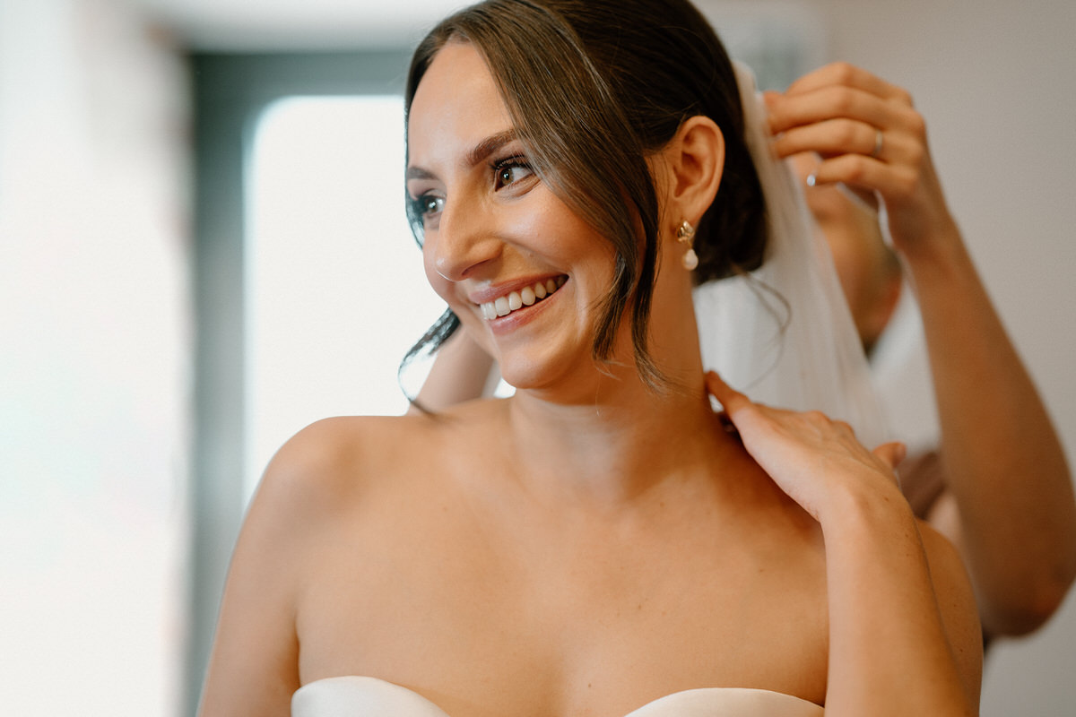 a beautiful bride smiles as a bridesmaid is helping with her finishing touches ahead of her wedding day