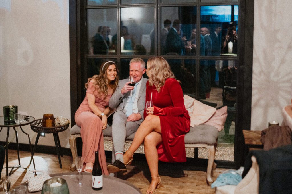 three wedding guests sat on a sofa chatting and drinking during a wedding reception at Botley Hill Barn as taken by a Luxury Wedding Photographer