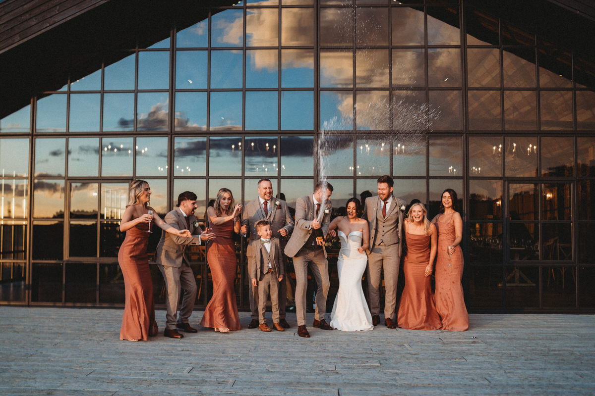 a bridal party celebrating a wedding by spraying champagne outside large barn windows at Botley Hill Barn