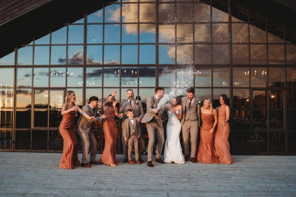 bridal party lined up spraying champagne in front of a wedding barn window at sunset