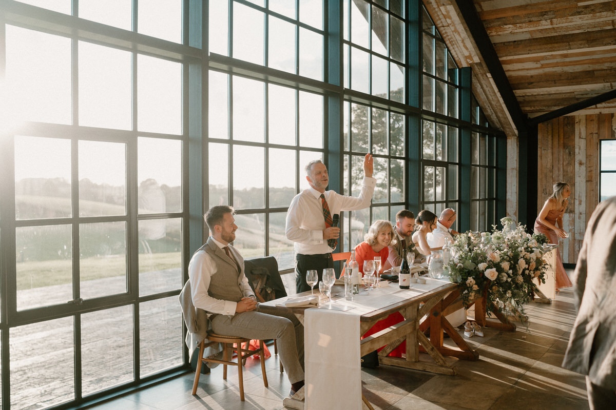 a man is stood giving a wedding speech at his sons wedding in a barn venue