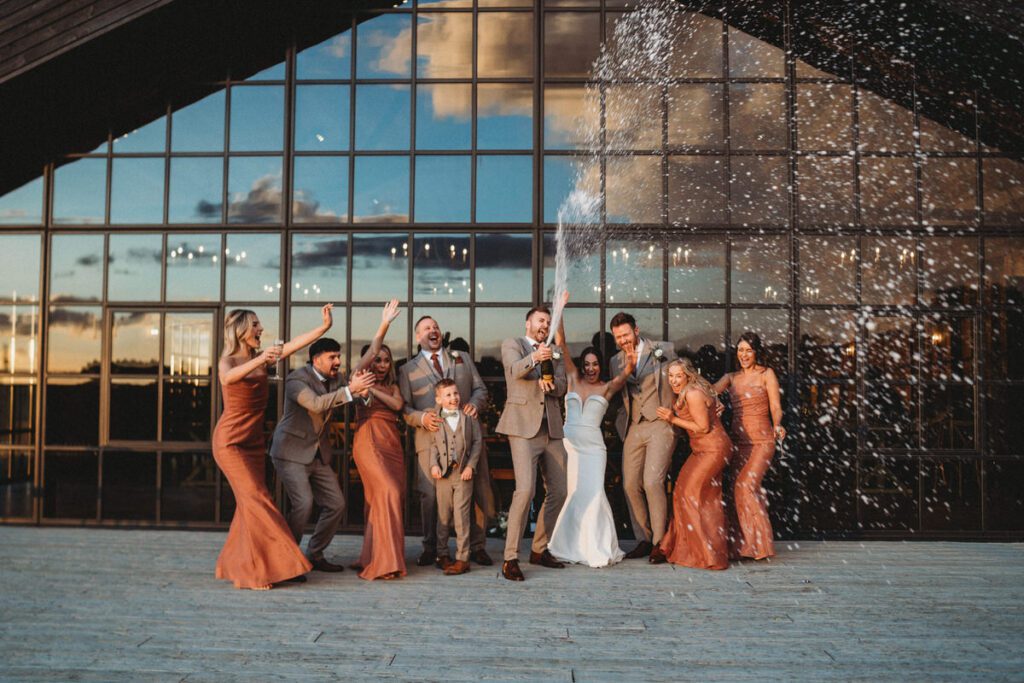 bridal party lined up spraying champagne in front of a wedding barn window at sunset