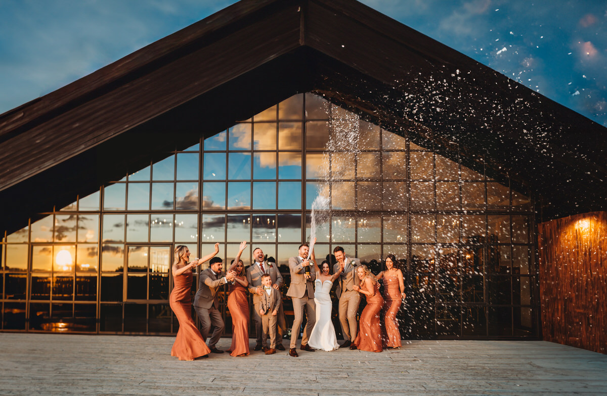 bridal party lined up spraying champagne in front of a wedding barn window at sunset