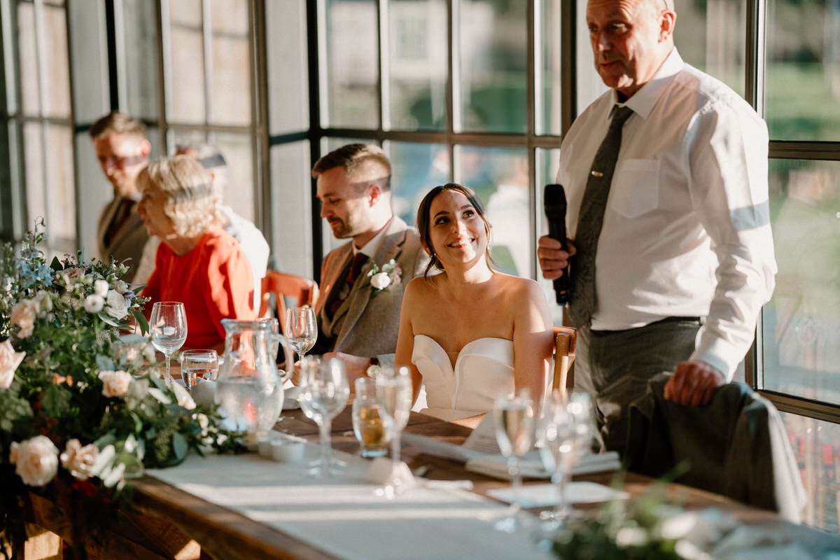 a bride looks on smiling as her father gives a wedding speech